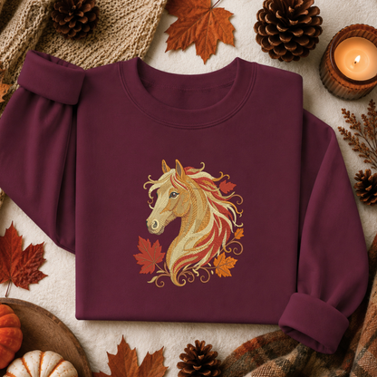 A folded long-sleeve crewneck sweatshirt in deep burgundy, featuring a detailed graphic of a golden horse head surrounded by autumn maple leaves. The sweatshirt is styled in a cozy flat-lay arrangement with pinecones, a lit candle, a knit scarf, and small pumpkins on a textured cream fabric background.