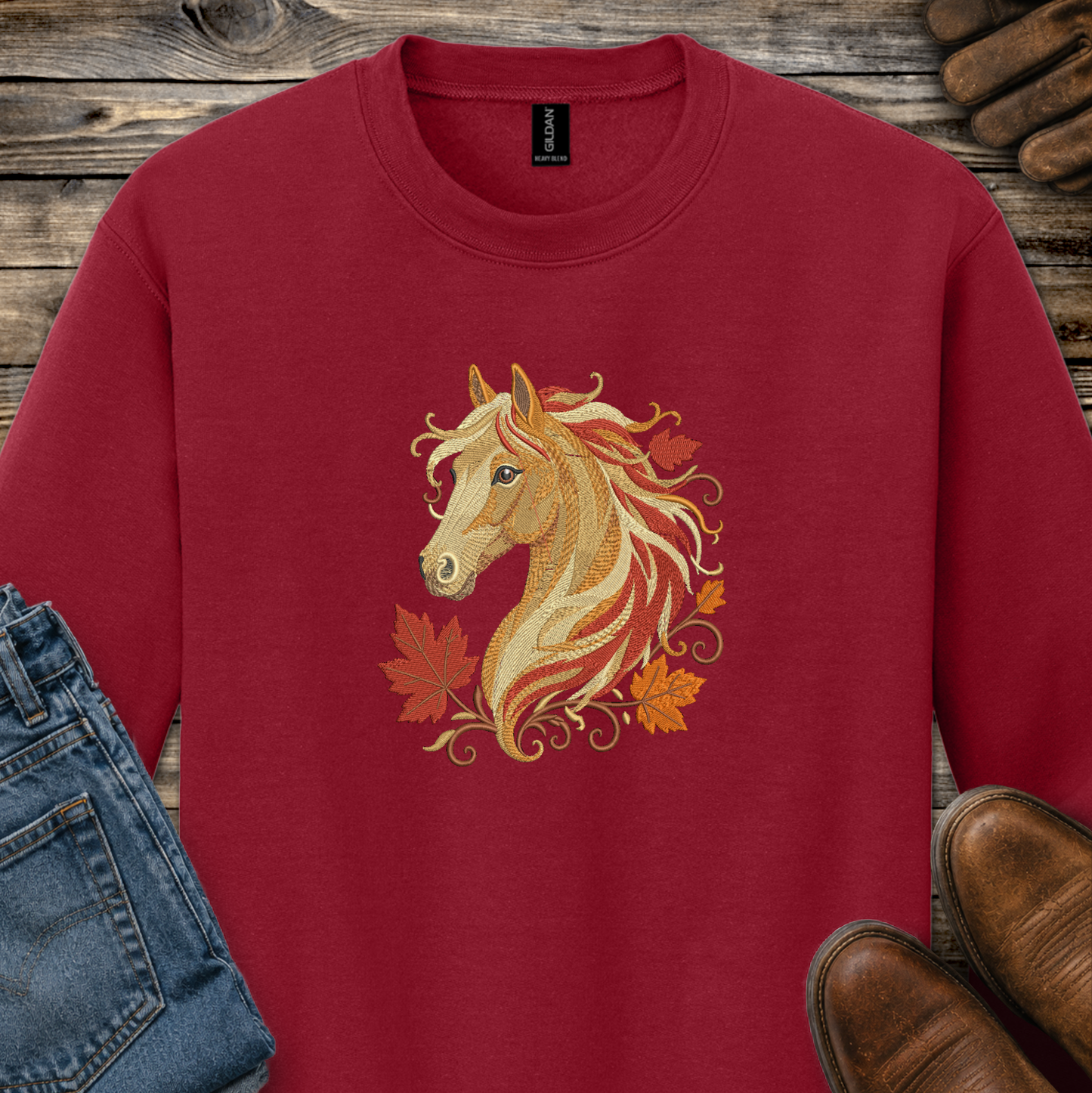 A red crewneck sweatshirt featuring a detailed graphic of a golden horse head surrounded by autumn leaves, styled in a flat-lay arrangement with jeans, brown shoes, and a rustic wood background.