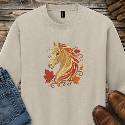 A beige crewneck sweatshirt featuring a graphic of a golden horse head surrounded by red and orange autumn leaves. The sweatshirt is styled in a flat-lay arrangement on a rustic wooden background, accompanied by a pair of blue jeans and brown leather shoes.