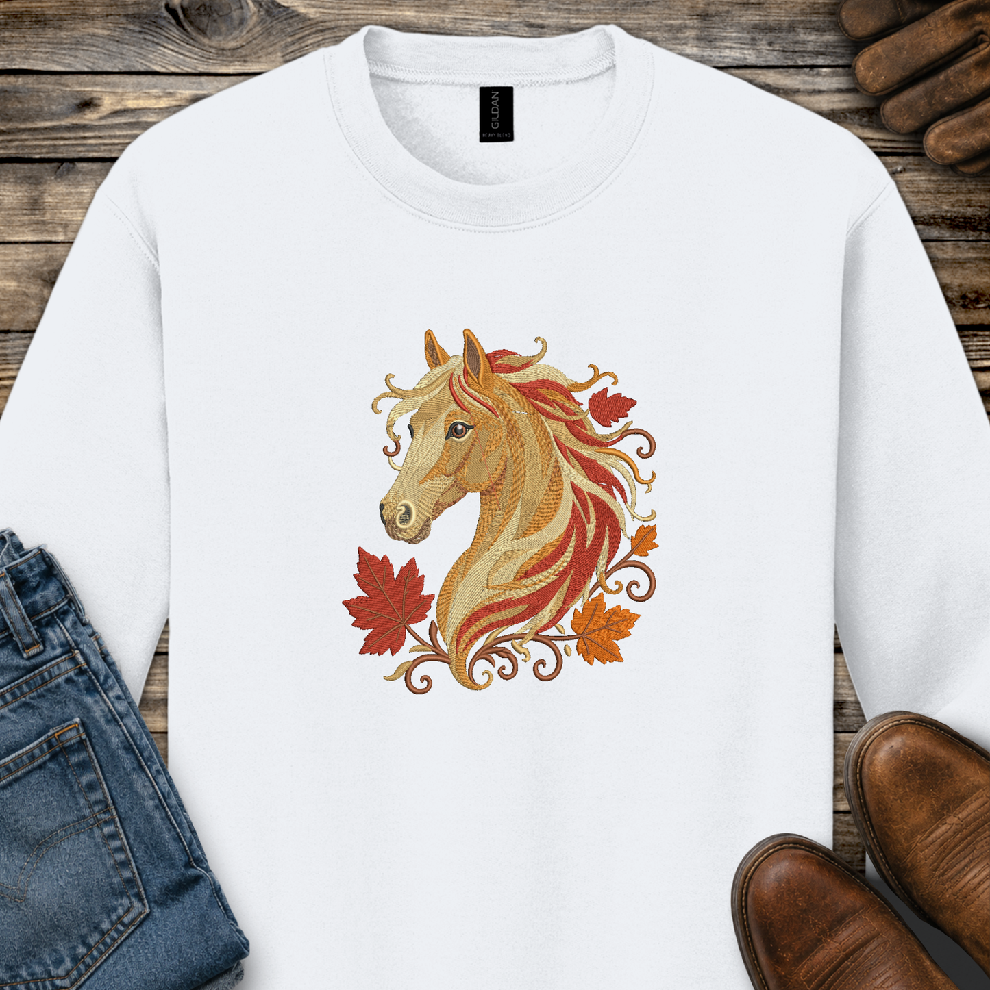 A white crewneck sweatshirt featuring a graphic of a golden horse head surrounded by red and orange autumn leaves, styled in a flat-lay on a rustic wooden background with jeans, shoes, and leather accessories.