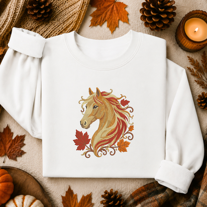 A white long-sleeve crewneck sweatshirt laid flat, featuring a central graphic of a stylized horse head with autumn leaves and swirls in tan, red, orange, and brown. The sweatshirt is styled in a cozy fall flat lay with pinecones, leaves, a candle, and plaid fabric.