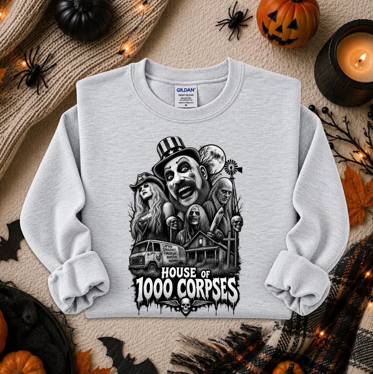 A light gray crewneck sweatshirt with a black and white graphic of horror movie villains and the text 'HOUSE OF 1000 CORPSES'. The sweatshirt is displayed in a flat-lay arrangement with Halloween props including pumpkins, a candle, spider decorations, and a knit sweater.