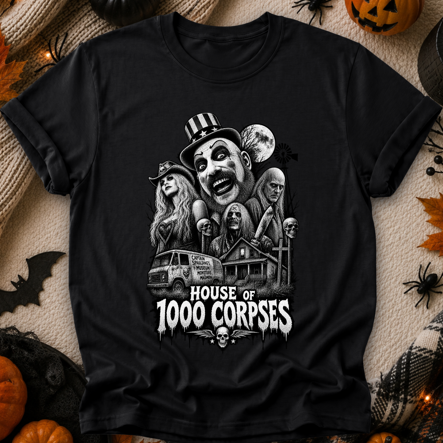 A black short-sleeve graphic T-shirt laid flat on a cozy beige fabric background with Halloween props including pumpkins, spider decorations, and a small bat. The shirt features a large black-and-white horror illustration of multiple faces and a haunted house, with the text 'HOUSE OF 1000 CORPSES' in white below.