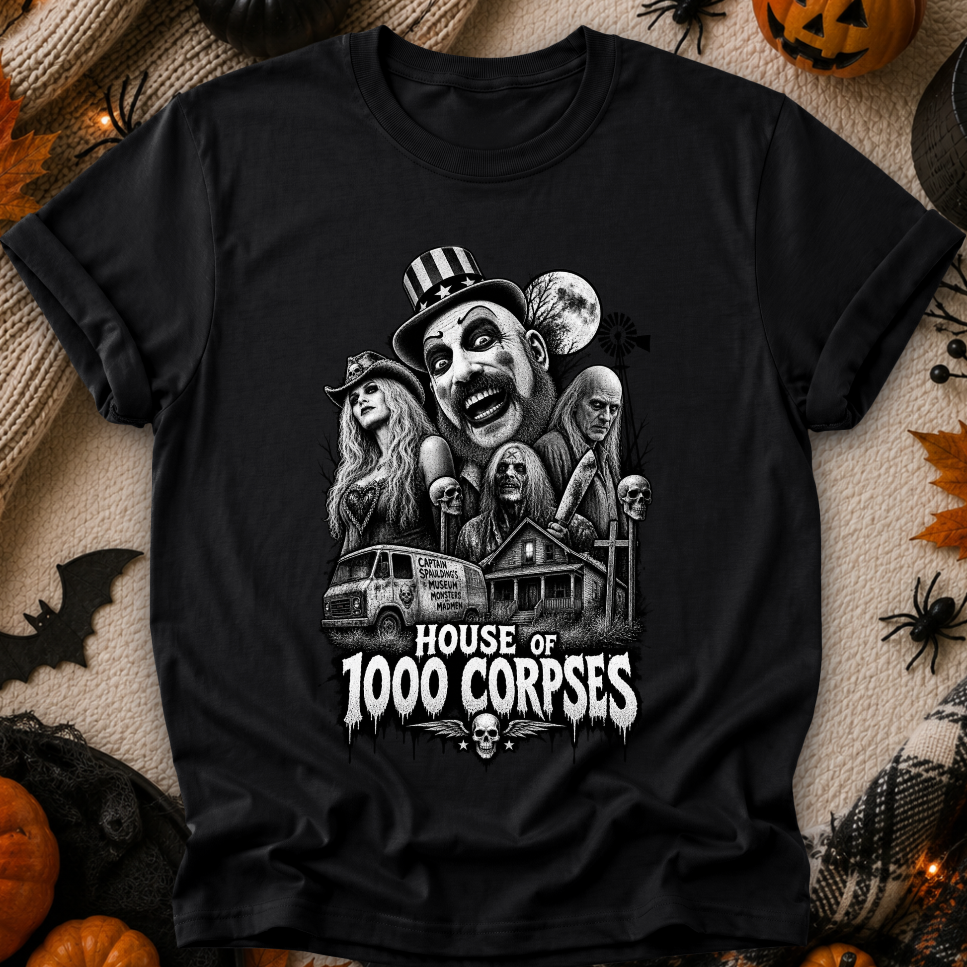 A black short-sleeve graphic T-shirt laid flat on a cozy beige fabric background with Halloween props including pumpkins, spider decorations, and a small bat. The shirt features a large black-and-white horror illustration of multiple faces and a haunted house, with the text 'HOUSE OF 1000 CORPSES' in white below.