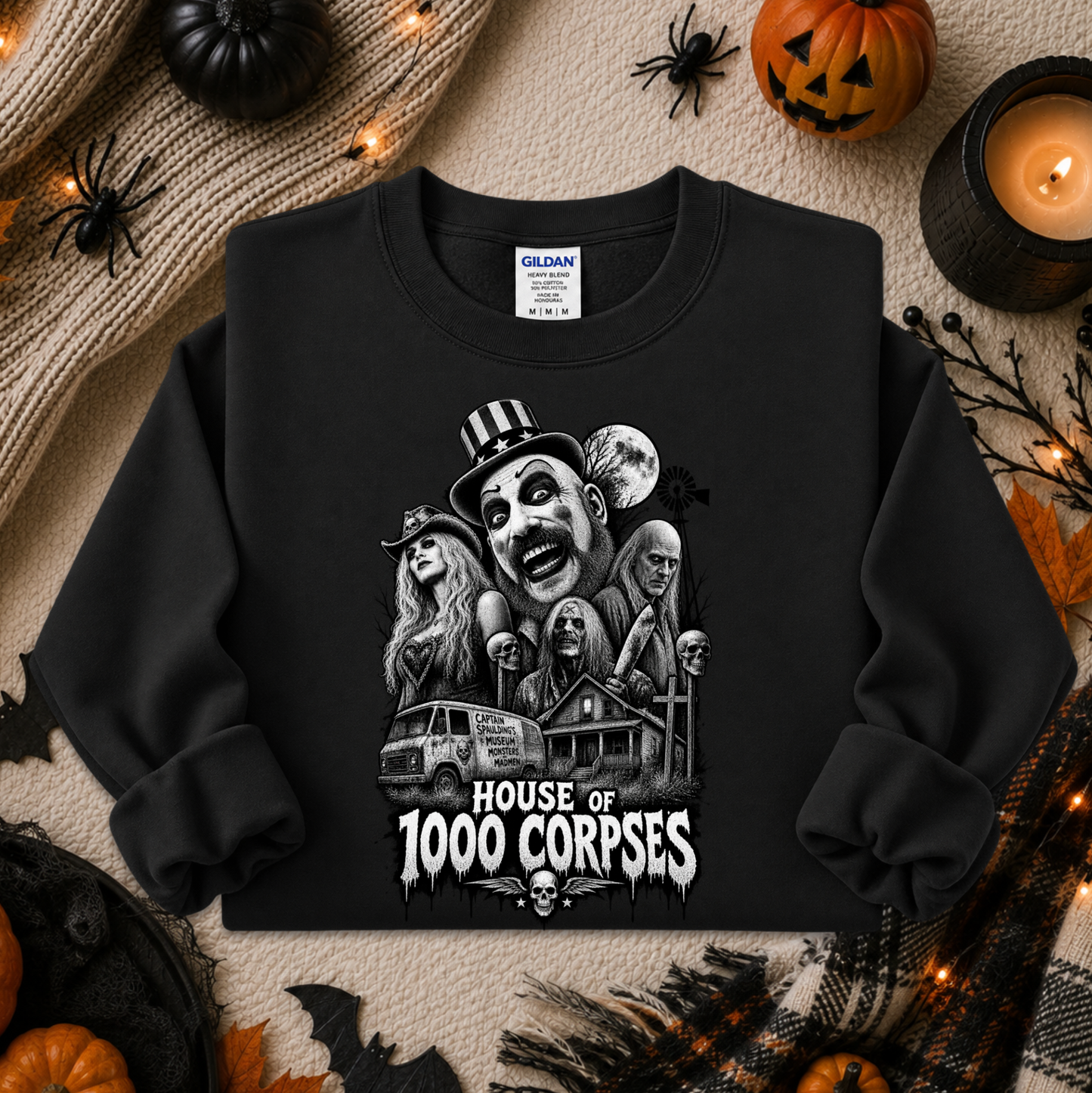 A black long-sleeve crewneck sweatshirt with a detailed black-and-white graphic of horror movie villains and the text 'HOUSE OF 1000 CORPSES' on the front. The sweatshirt is displayed in a flat-lay Halloween setting with pumpkins, a candle, spider decorations, and a knit sweater.