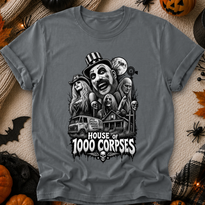 A dark gray graphic t-shirt featuring a black and white horror illustration of 'The House of 1000 Corpses' with a clown, ghosts, a van, and a haunted house, displayed on a cozy autumn-themed background with pumpkins, a knitted blanket, and spider decorations.