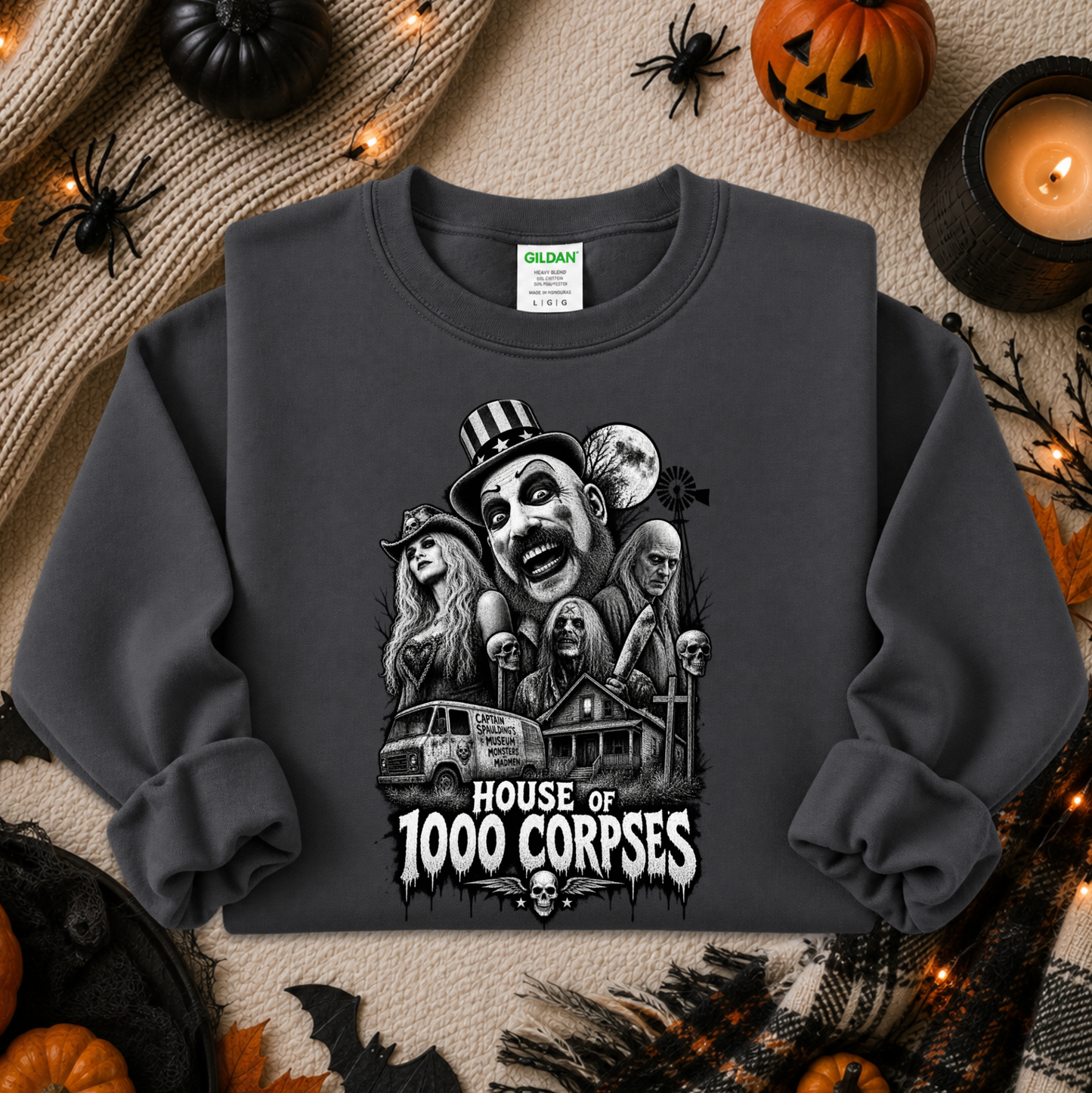 A dark gray crewneck sweatshirt with a detailed black and white graphic of horror movie characters and the text 'HOUSE OF 1000 CORPSES'. The sweatshirt is displayed in a flat-lay Halloween setting with pumpkins, a candle, and spider decorations.