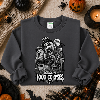 A dark gray crewneck sweatshirt with a detailed black and white graphic of horror movie characters and the text 'HOUSE OF 1000 CORPSES'. The sweatshirt is displayed in a flat-lay Halloween setting with pumpkins, a candle, and spider decorations.
