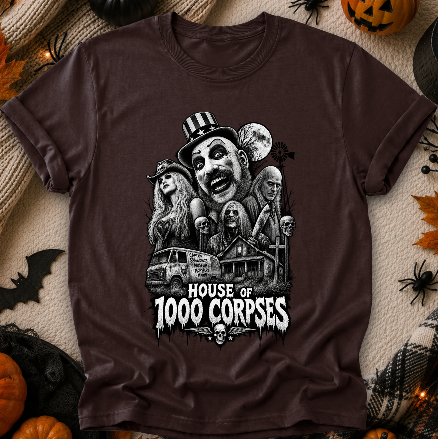 A dark brown short-sleeve graphic t-shirt laid flat on a cozy beige blanket surrounded by Halloween decorations, including pumpkins, a spider, a bat, and a jack-o'-lantern. The shirt features a detailed black and white graphic of a creepy clown-like character, a witch, a ghost, a van, a haunted house, and the text 'HOUSE OF 1000 CORPSES'.