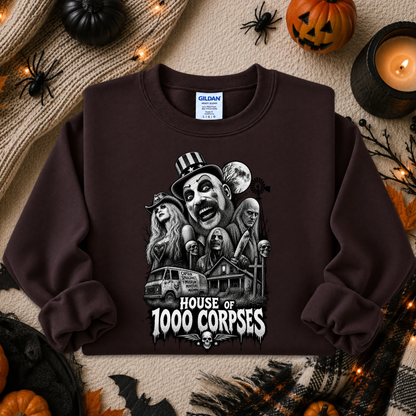 A dark brown crewneck sweatshirt featuring a black and white graphic illustration of the 'House of 1000 Corpses' characters, styled in a cozy autumn flat-lay with pumpkins, a candle, and a knitted blanket.