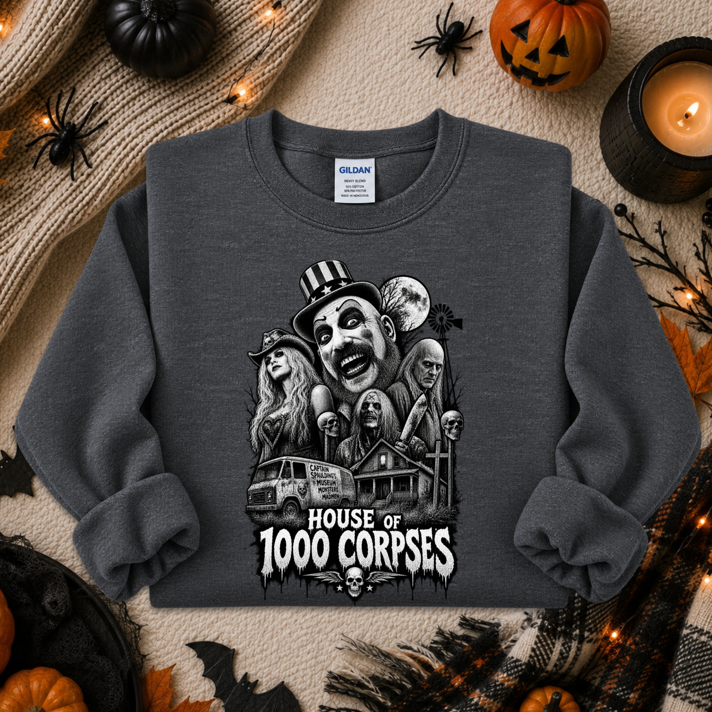 A dark gray crewneck sweatshirt featuring a black and white graphic of horror movie characters and the text 'HOUSE OF 1000 CORPSES'. The sweatshirt is displayed in a flat-lay arrangement with Halloween decorations including a pumpkin, spider, candle, and knitted blanket.
