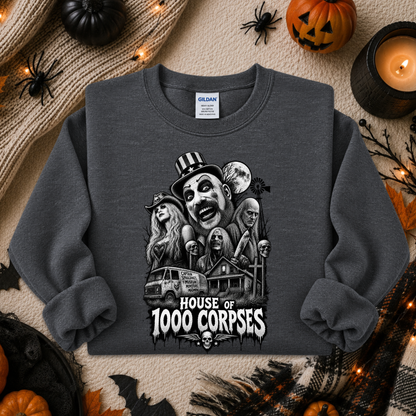 A dark gray crewneck sweatshirt featuring a black and white graphic of horror movie characters and the text 'HOUSE OF 1000 CORPSES'. The sweatshirt is displayed in a flat-lay arrangement with Halloween decorations including a pumpkin, spider, candle, and knitted blanket.