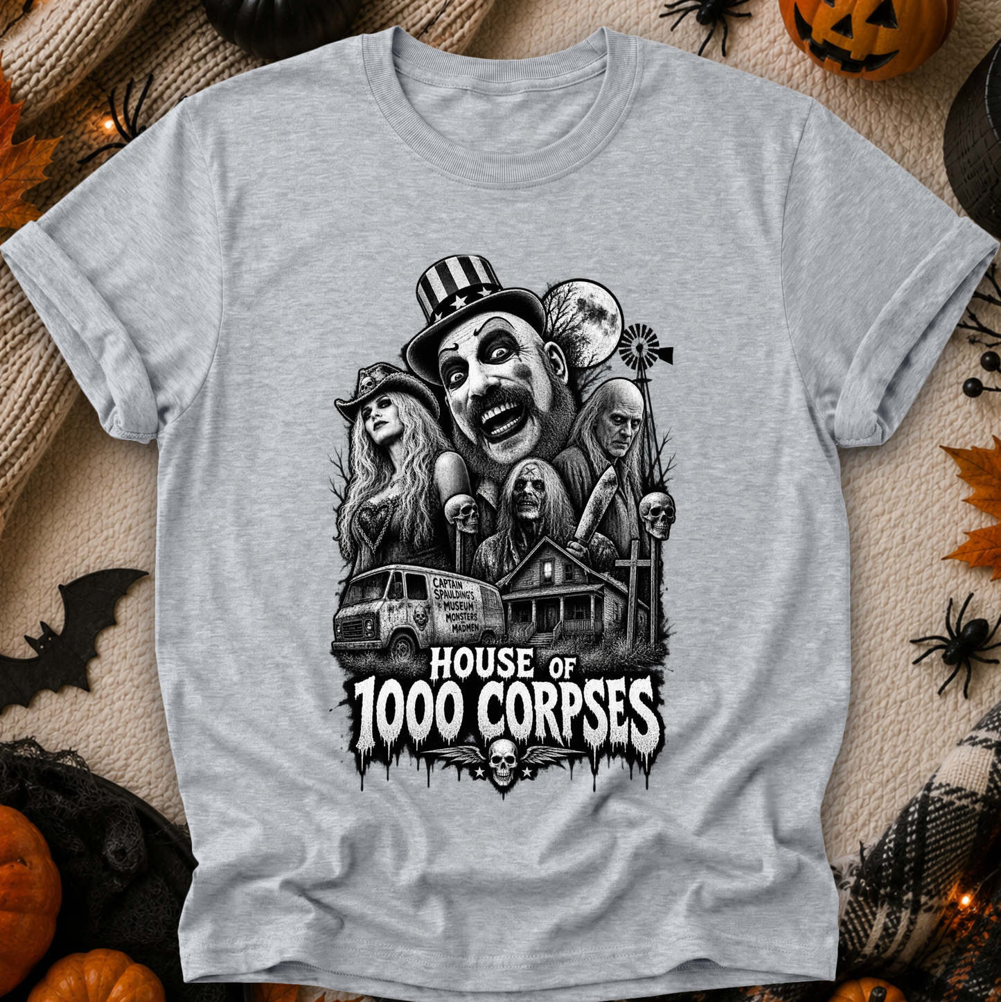A heather gray short-sleeve t-shirt with a black and white horror graphic print reading 'HOUSE OF 1000 CORPSES' and featuring illustrated faces, a van, a house, and skulls. The shirt is laid flat on a textured beige fabric surrounded by Halloween props including pumpkins, spiderwebs, and a bat.
