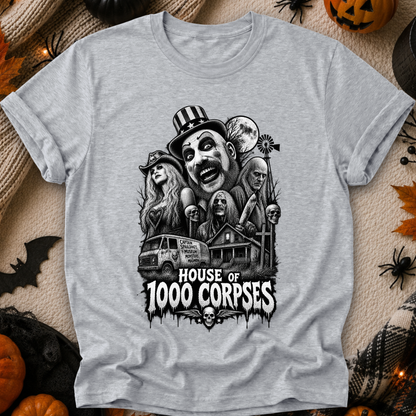 A heather gray short-sleeve t-shirt with a black and white horror graphic print reading 'HOUSE OF 1000 CORPSES' and featuring illustrated faces, a van, a house, and skulls. The shirt is laid flat on a textured beige fabric surrounded by Halloween props including pumpkins, spiderwebs, and a bat.