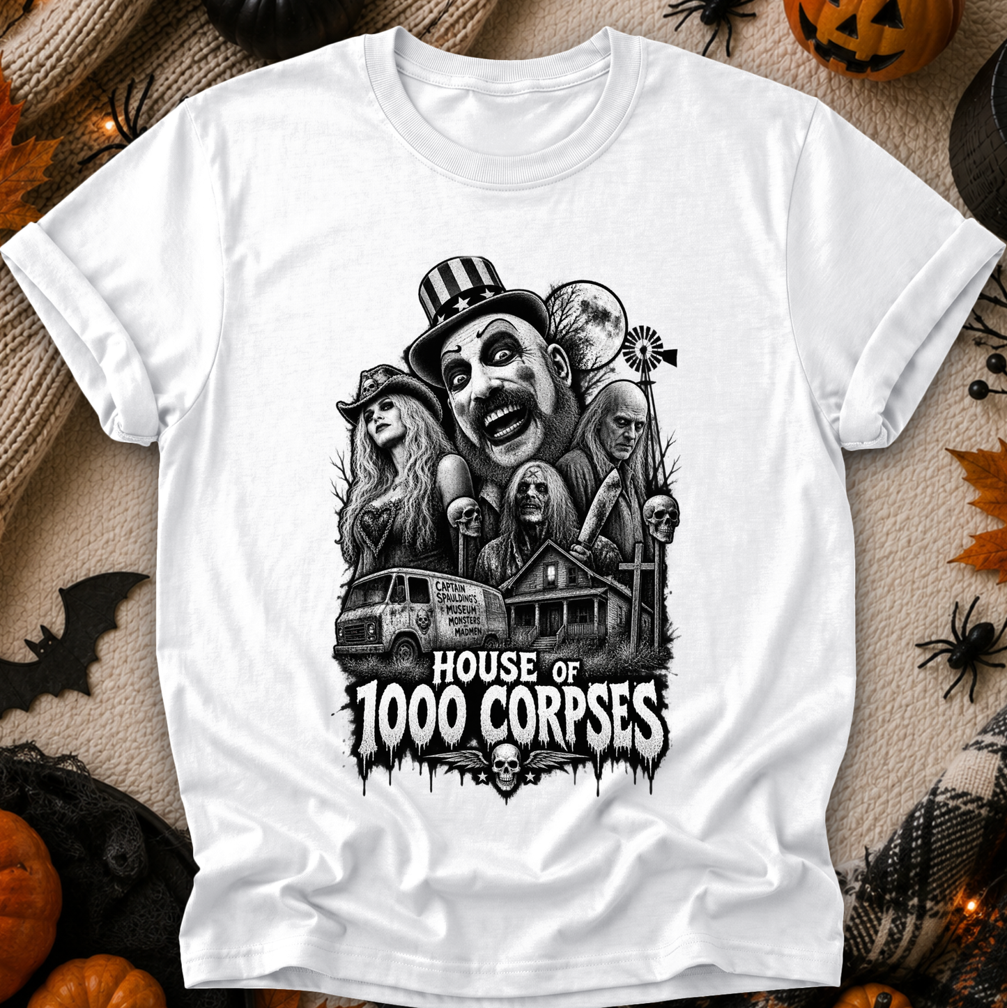 A white short-sleeve graphic T-shirt laid flat on a Halloween-themed background with pumpkins, spiders, and black fabric decor. The shirt features a large black-and-white horror illustration of multiple faces, a van, a haunted house, and the text 'HOUSE OF 1000 CORPSES.