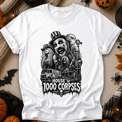 A white short-sleeve graphic T-shirt laid flat on a Halloween-themed background with pumpkins, spiders, and black fabric decor. The shirt features a large black-and-white horror illustration of multiple faces, a van, a haunted house, and the text 'HOUSE OF 1000 CORPSES.