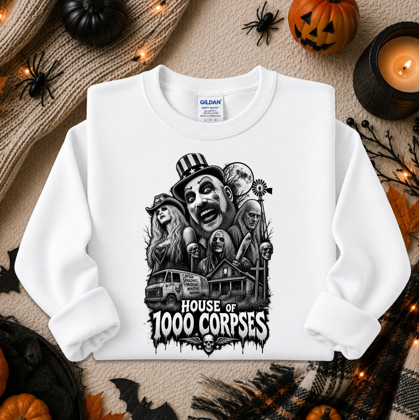A white long-sleeve crewneck sweatshirt with a black graphic print of horror characters and the text 'HOUSE OF 1000 CORPSES'. The sweatshirt is displayed in a flat-lay Halloween setting with pumpkins, a candle, and a knitted blanket.