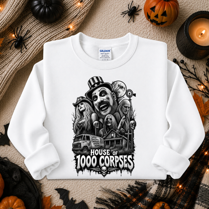 A white long-sleeve crewneck sweatshirt with a black graphic print of horror characters and the text 'HOUSE OF 1000 CORPSES'. The sweatshirt is displayed in a flat-lay Halloween setting with pumpkins, a candle, and a knitted blanket.