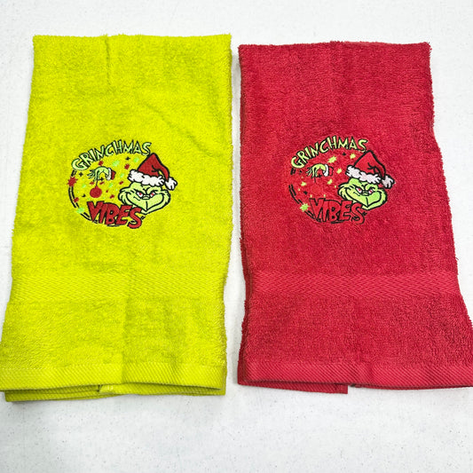Two folded towels side by side on a white background, one bright yellow-green and one red, both featuring a Grinch-themed holiday graphic with the text 'Grinchmas Vibes'.