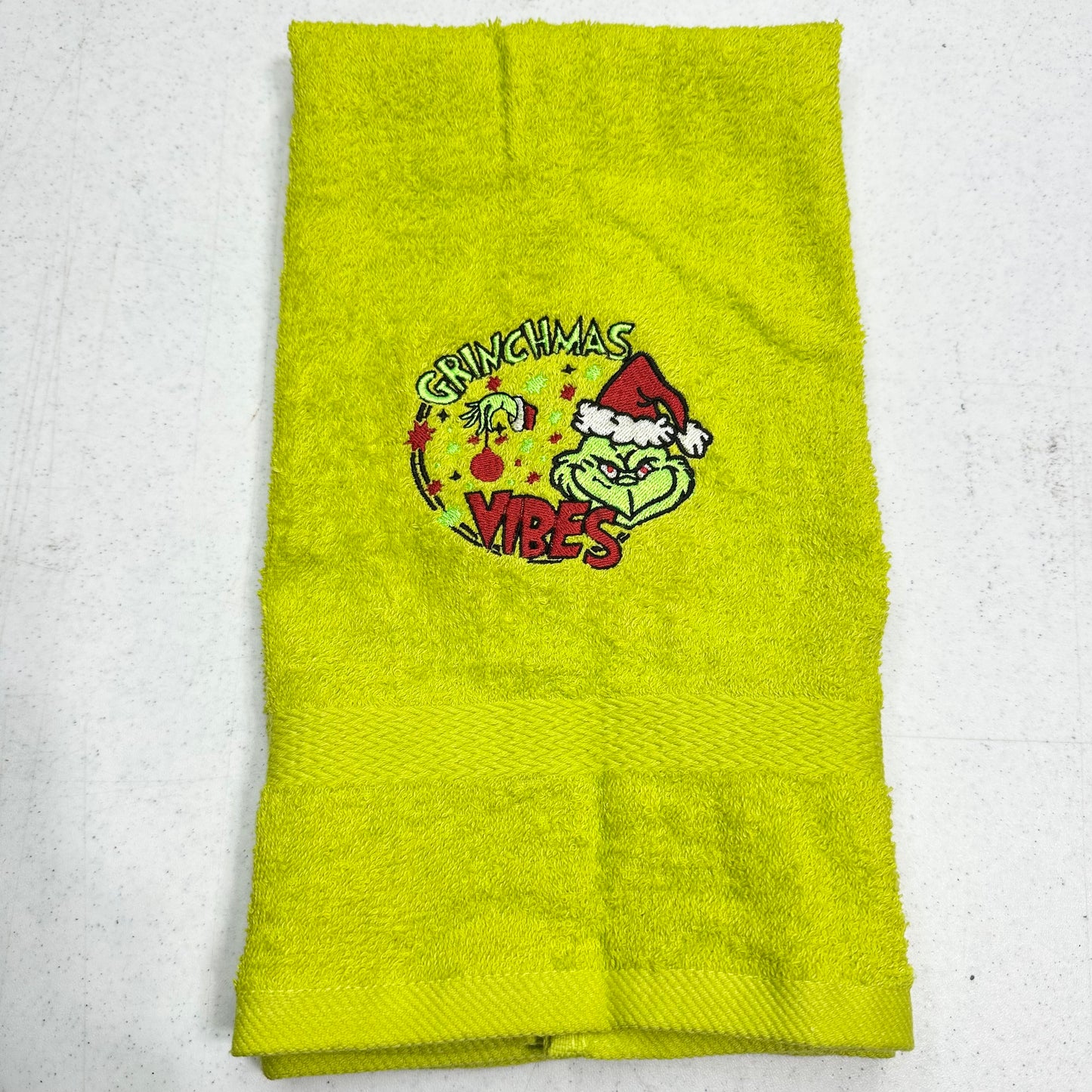 A lime green towel featuring a Grinch-themed Christmas graphic with the words 'Grinchmas Vibes' and a cartoon Grinch wearing a Santa hat, laid flat on a light gray surface.