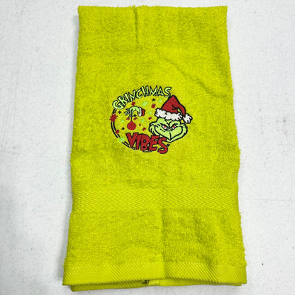 A lime green towel featuring a Grinch-themed Christmas graphic with the words 'Grinchmas Vibes' and a cartoon Grinch wearing a Santa hat, laid flat on a light gray surface.