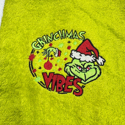 A close-up of a bright lime green plush towel featuring an embroidered Grinch-themed design with the words 'Grinchmas Vibes' and a Santa hat.