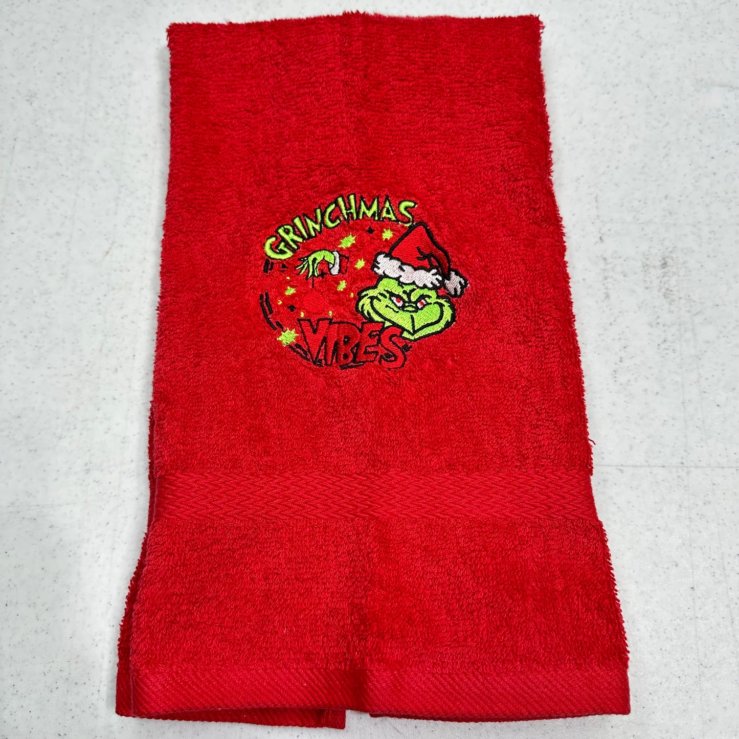 A red terry cloth hand towel featuring a Grinch-themed embroidered graphic in the center with the words 'GRINCHMAS IT'S VIBES' and a cartoon Grinch wearing a Santa hat.