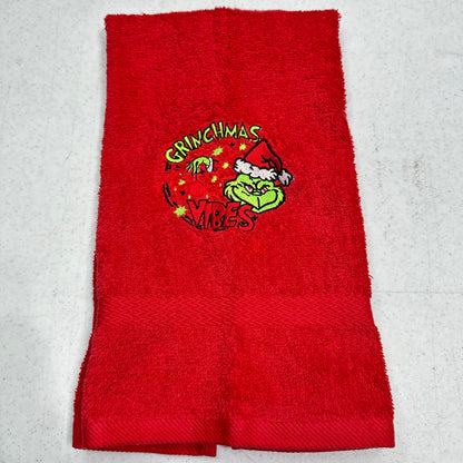 A red terry cloth hand towel featuring a Grinch-themed embroidered graphic in the center with the words 'GRINCHMAS IT'S VIBES' and a cartoon Grinch wearing a Santa hat.