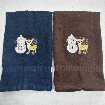 Two folded towels side by side on a light gray surface. The left towel is navy blue and the right towel is brown, both featuring an embroidered winter design of a snowman and a yellow deer.