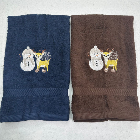 Two folded towels side by side on a light gray surface. The left towel is navy blue and the right towel is brown, both featuring an embroidered winter design of a snowman and a yellow deer.