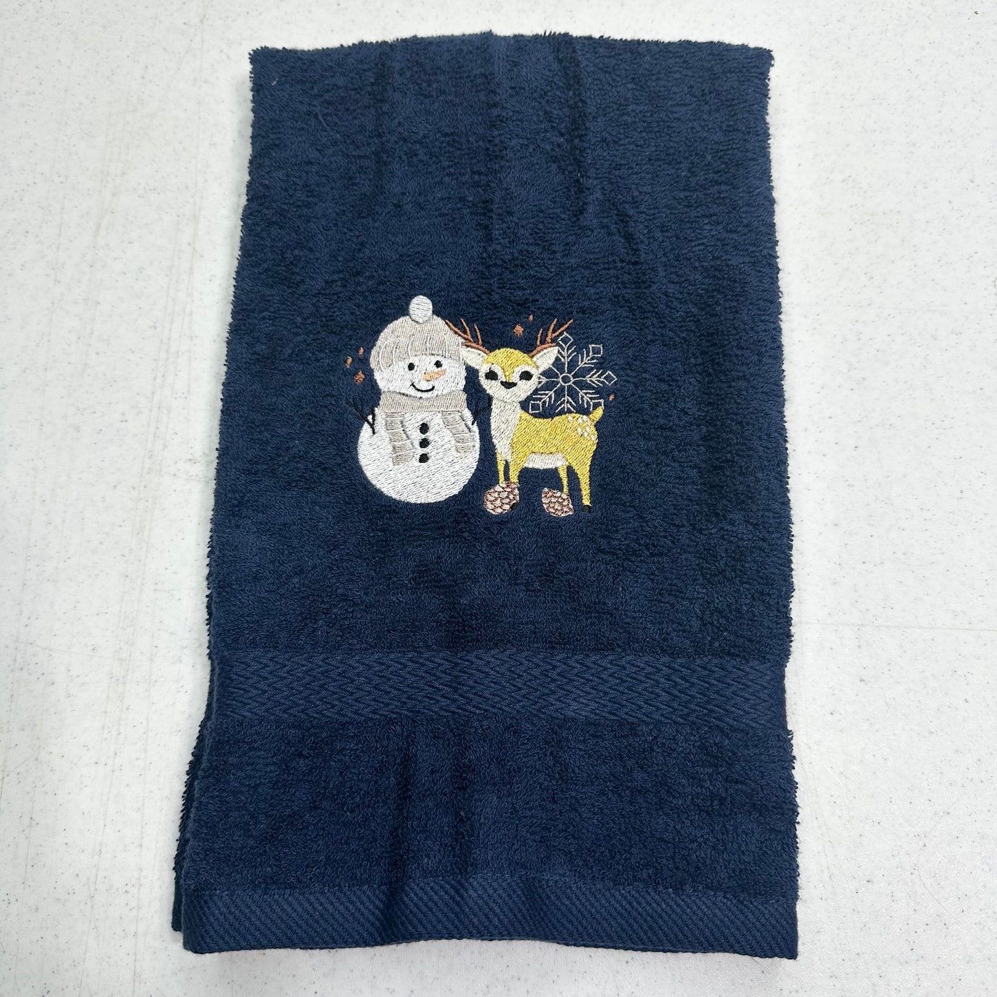 A dark navy blue towel with a decorative embroidered design of a white snowman and a yellow/gold deer, laid flat on a light gray textured surface.