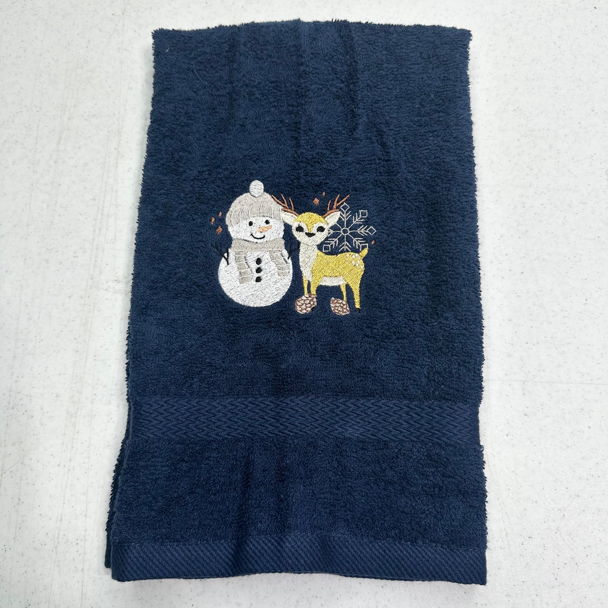 A dark navy blue towel with a decorative embroidered design of a white snowman and a yellow/gold deer, laid flat on a light gray textured surface.