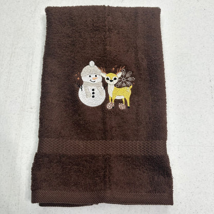 A dark brown terry cloth hand towel featuring an embroidered design of a white snowman and a yellow deer with antlers on the front, laid flat on a light gray textured surface.