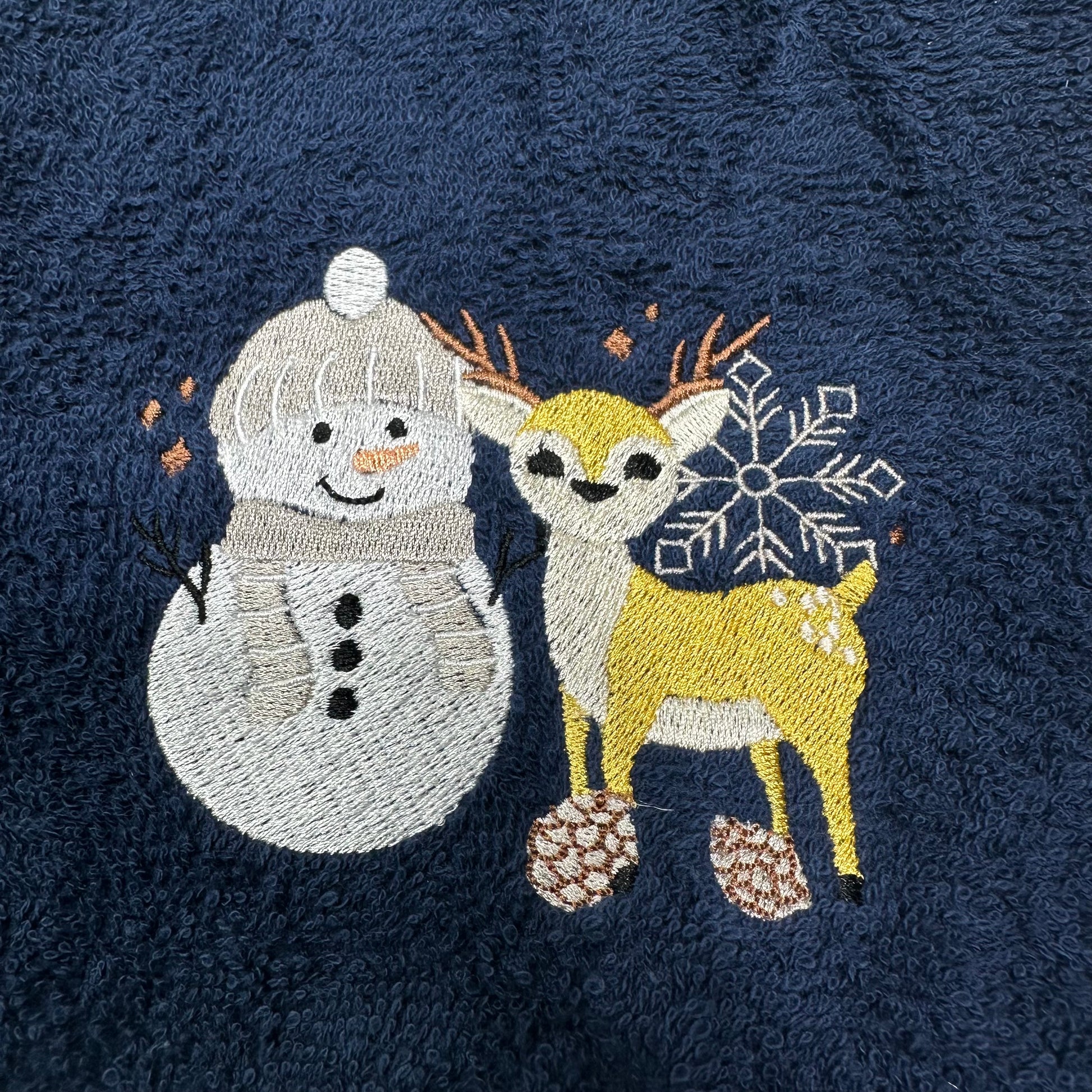 A close-up of a dark navy blue textured fabric, likely a towel or blanket, featuring embroidered winter motifs of a white snowman and a yellow deer with a white snowflake and pinecones.