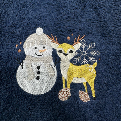 A close-up of a dark navy blue textured fabric, likely a towel or blanket, featuring embroidered winter motifs of a white snowman and a yellow deer with a white snowflake and pinecones.