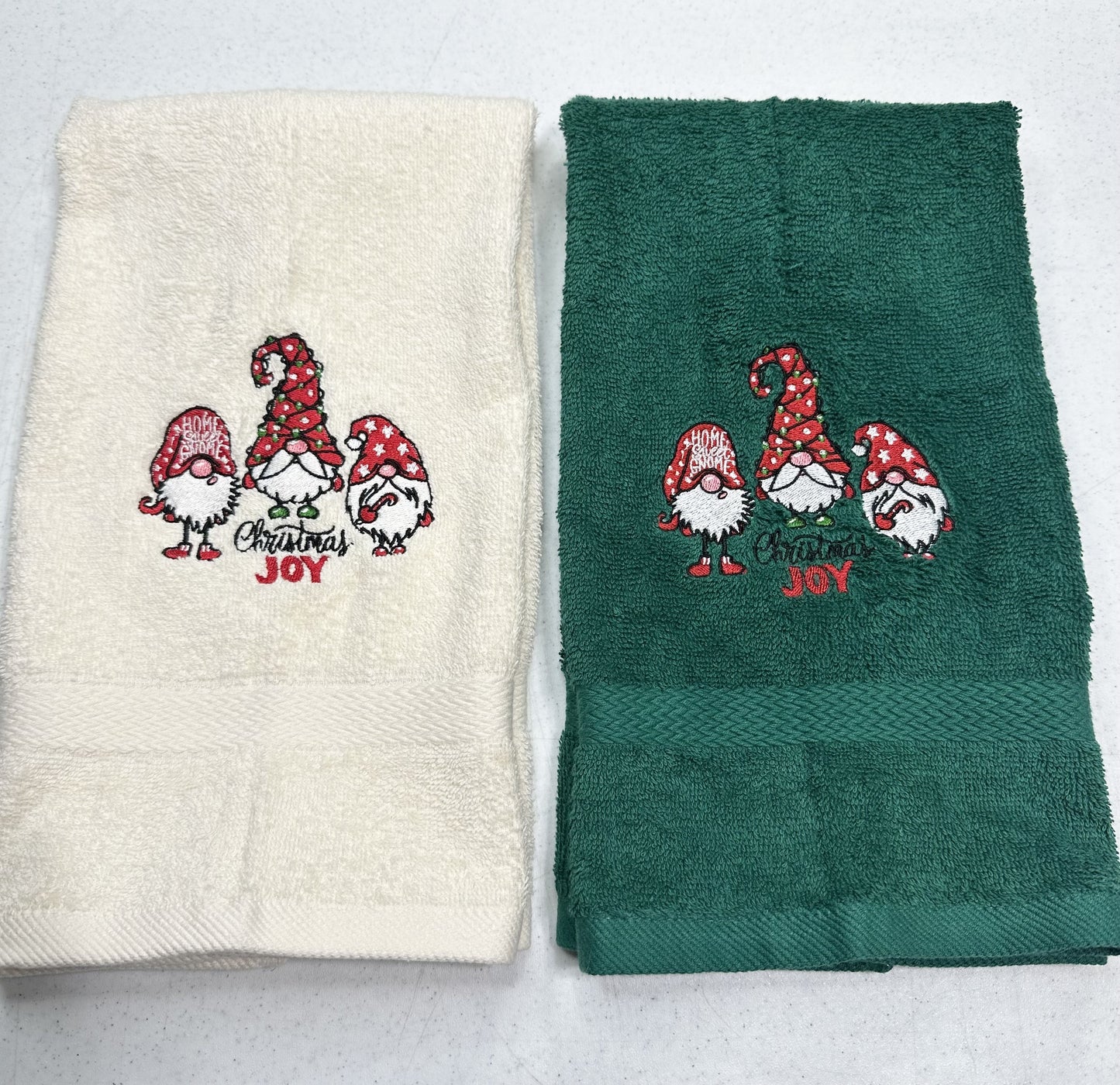 Two folded towels side by side on a light gray background. The left towel is cream with a red and green Christmas gnome embroidery that reads 'Merry Christmas Joy'.