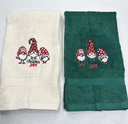 Two folded towels side by side on a light gray background. The left towel is cream with a red and green Christmas gnome embroidery that reads 'Merry Christmas Joy'.