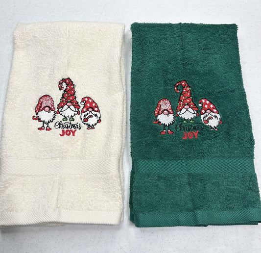 Two folded towels side by side on a light gray background. The left towel is cream with a red and green Christmas gnome embroidery that reads 'Merry Christmas Joy'.
