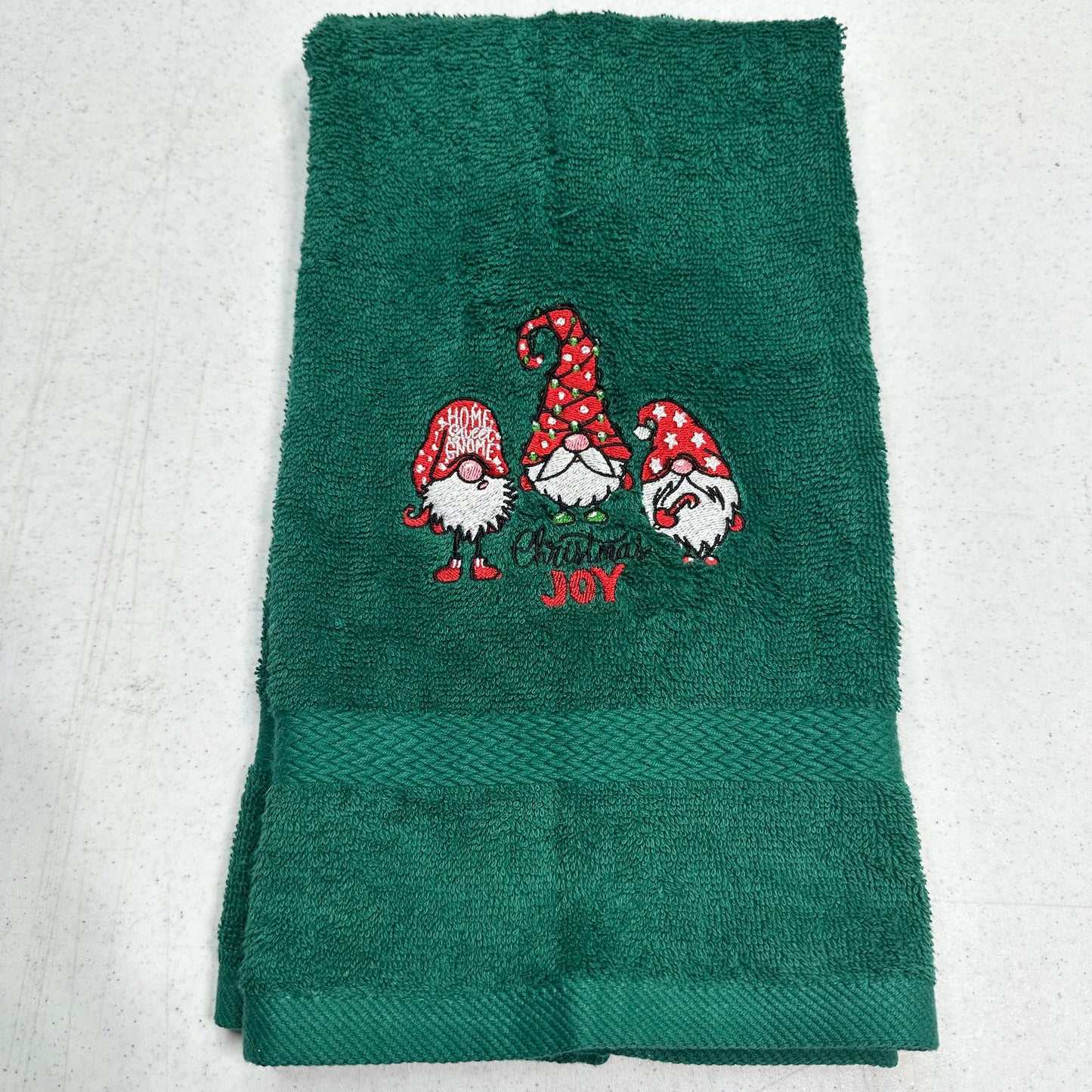 A dark green terry cloth towel featuring three embroidered Christmas garden gnomes and the words 'MERRY JOY' in red and white embroidery, folded on a light gray textured background.