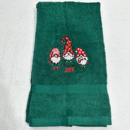 A dark green terry cloth towel featuring three embroidered Christmas garden gnomes and the words 'MERRY JOY' in red and white embroidery, folded on a light gray textured background.