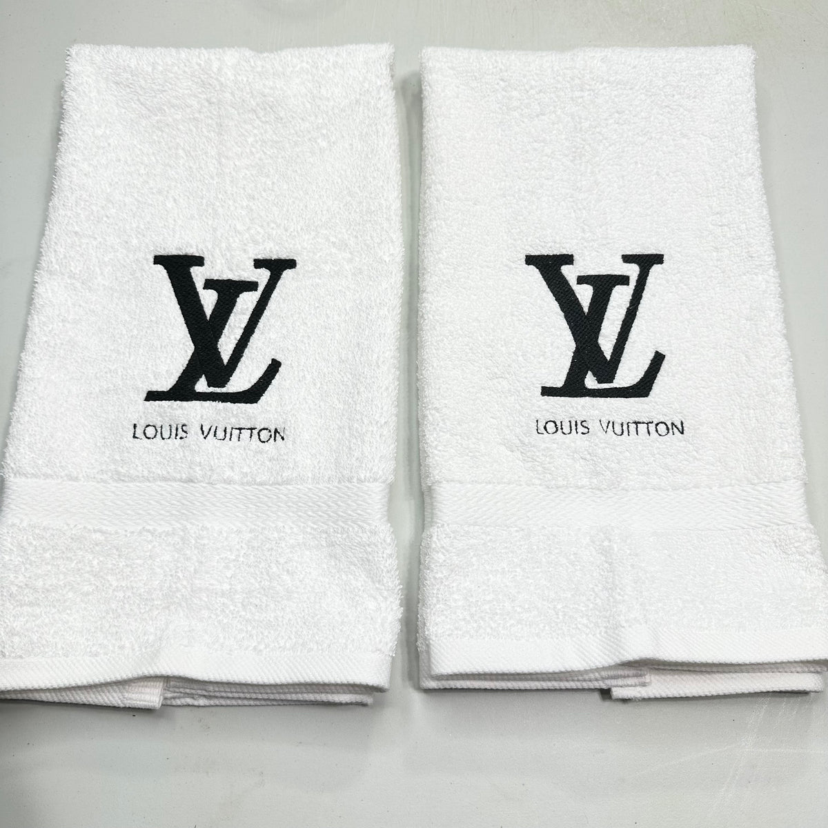 Two folded white cotton hand towels with black embroidered Louis Vuitton LV monogram and 'LOUIS VUITTON' text on the front, displayed side by side on a light gray surface.