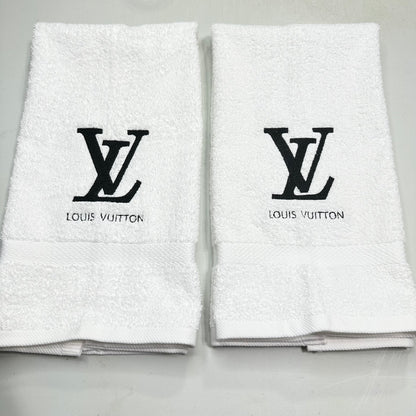 Two folded white cotton hand towels with black embroidered Louis Vuitton LV monogram and 'LOUIS VUITTON' text on the front, displayed side by side on a light gray surface.