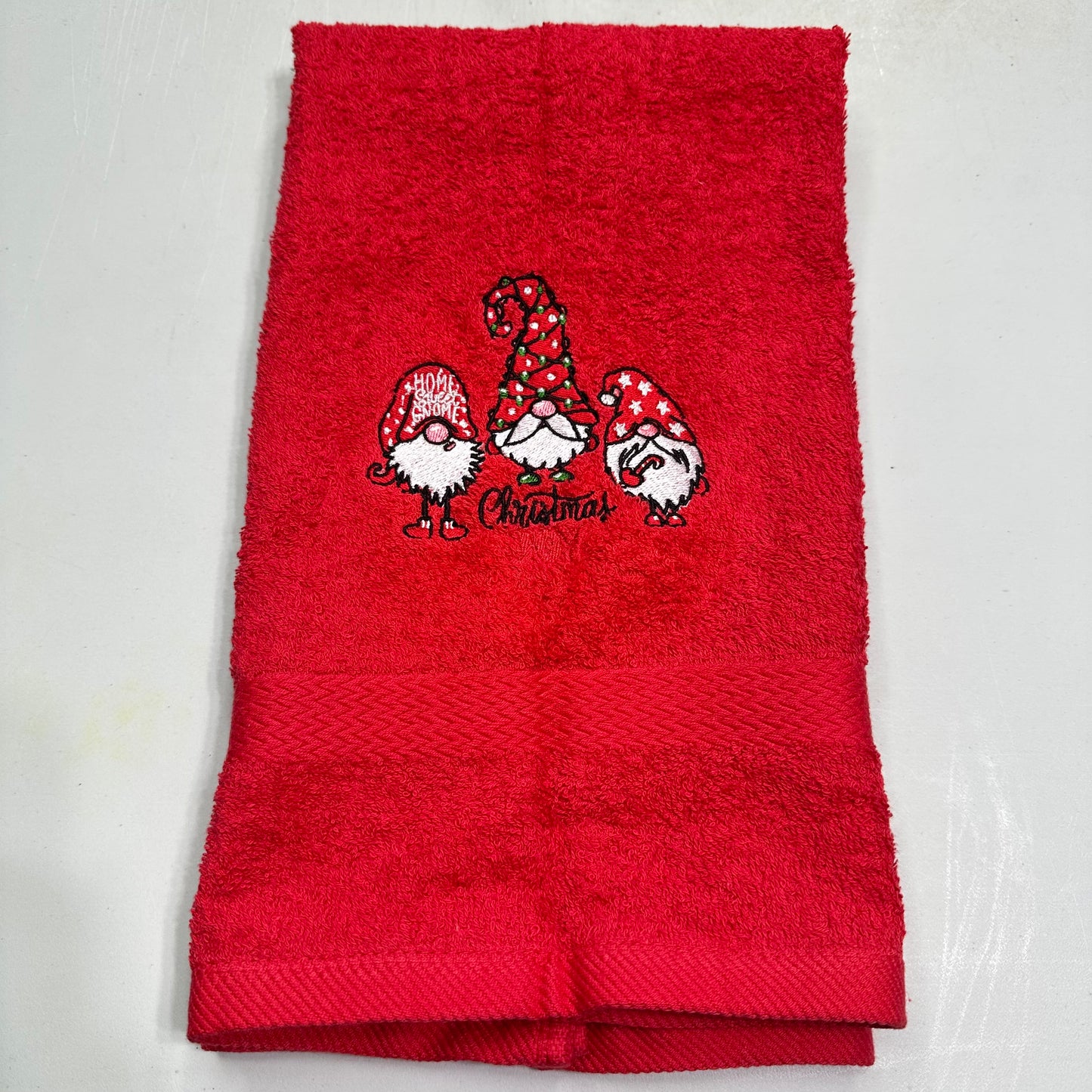 A folded red terry cloth towel featuring an embroidered design of three Christmas gnomes with red and white patterned hats and the word 'Christmas' stitched below them, shown on a light gray background.