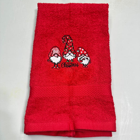 A folded red terry cloth towel featuring an embroidered design of three Christmas gnomes with red and white patterned hats and the word 'Christmas' stitched below them, shown on a light gray background.