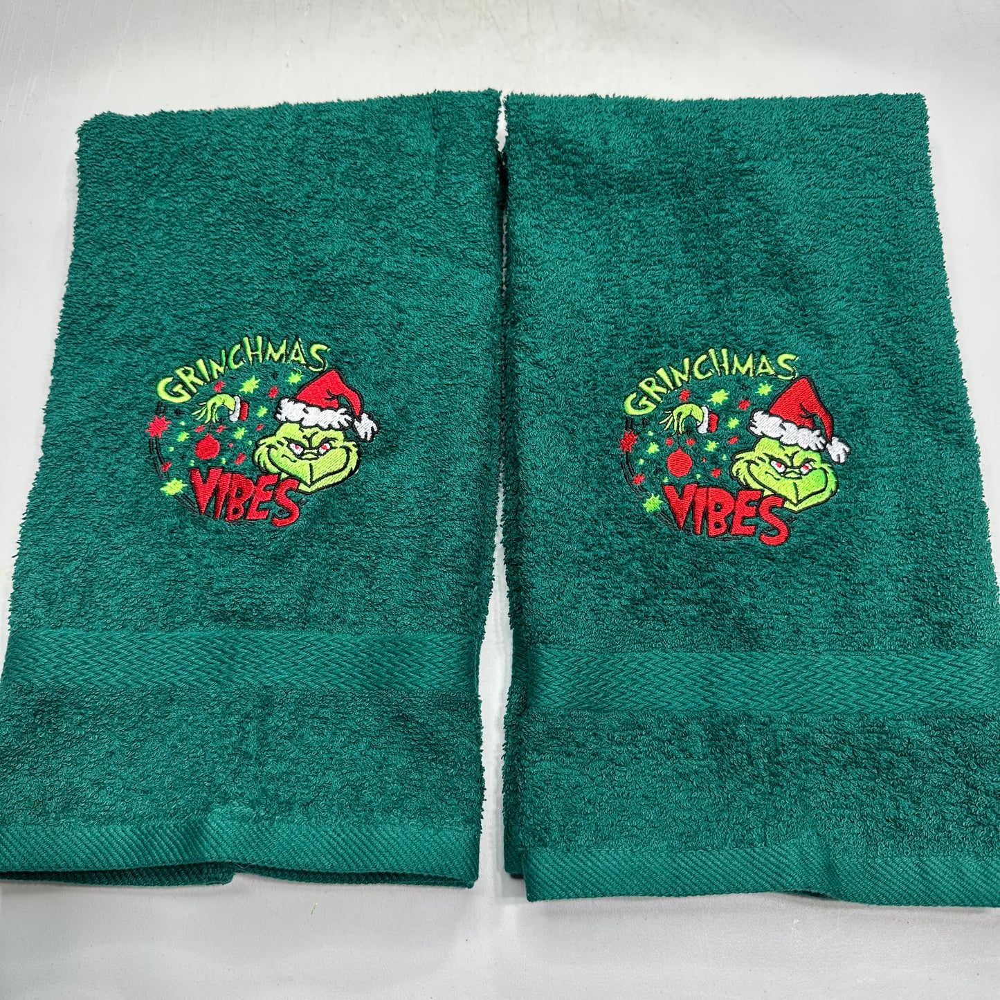 Two dark green towels with embroidered Grinch and Christmas graphics, displayed side-by-side on a white background.