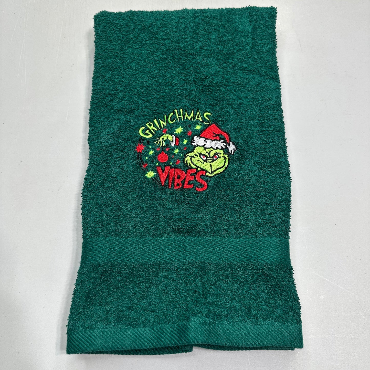 A dark green terry cloth hand towel featuring an embroidered Grinch graphic with the words 'Grinchmas Vibes' and a Santa hat, laid flat on a light gray background.