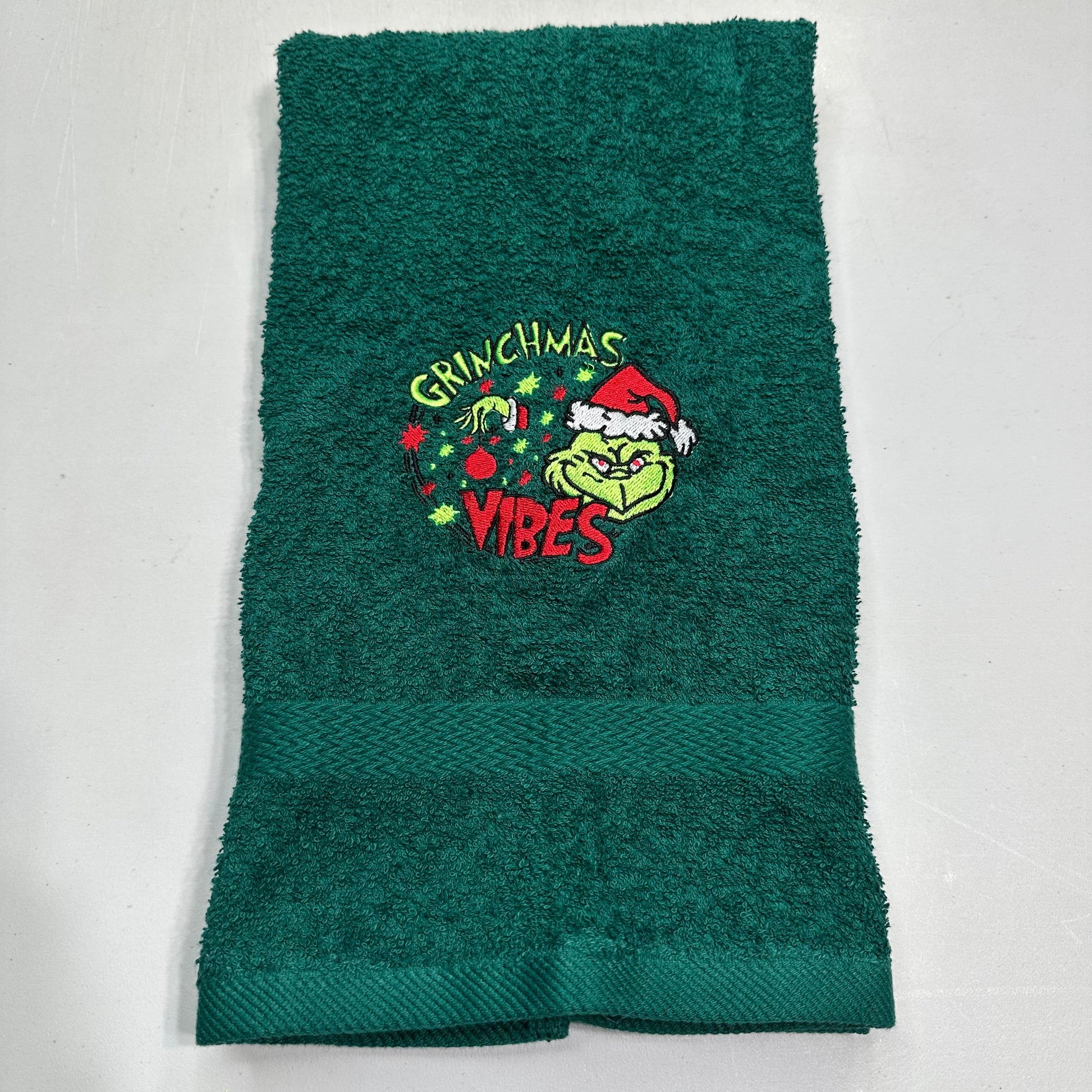 A dark green terry cloth hand towel featuring an embroidered Grinch graphic with the words 'Grinchmas Vibes' and a Santa hat, laid flat on a light gray background.