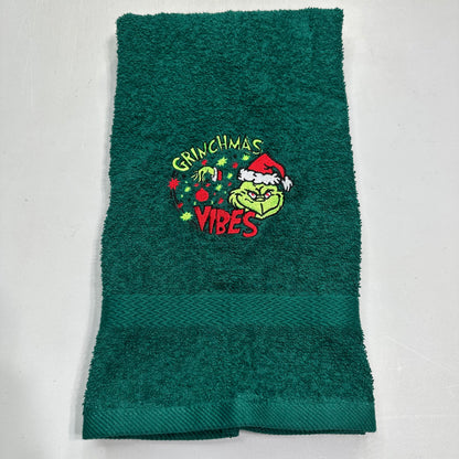 A dark green terry cloth hand towel featuring an embroidered Grinch graphic with the words 'Grinchmas Vibes' and a Santa hat, laid flat on a light gray background.