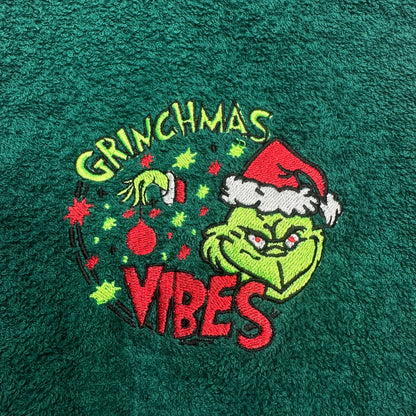 A close-up of a dark green textured fabric featuring an embroidered Grinch graphic with the words 'GRINCHMAS VIBES' and a Christmas ornament.