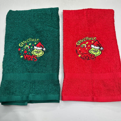 Two folded terry cloth towels side by side on a white background, one dark green and one red, both featuring an embroidered Grinch-themed holiday design with the words 'Grinchmas Vibes'.